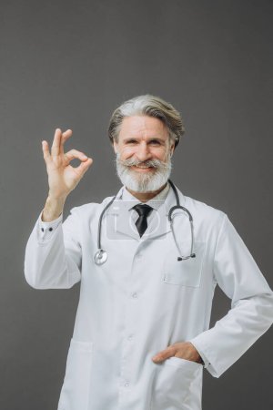 Photo for Gray-haired middle-aged man in doctor's coat with stethoscope, showing ok gesture, isolated on gray background. - Royalty Free Image