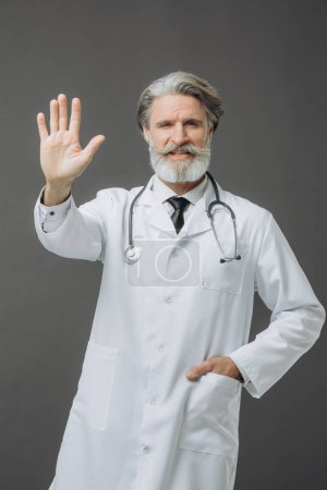 Photo for Gray-haired male doctor in white medical coat giving high five, isolated on gray background. - Royalty Free Image