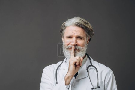 Photo for Gray-haired male doctor in white medical coat isolated on gray background raising hand to mouth with silence gesture - Royalty Free Image