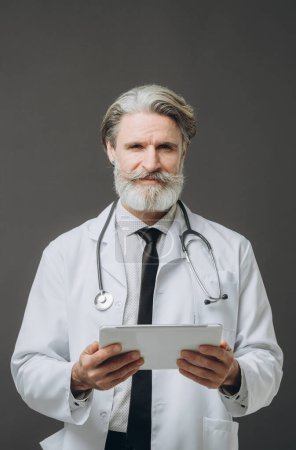 Photo for Gray-haired handsome male doctor with a tablet in his hands on a gray background. - Royalty Free Image