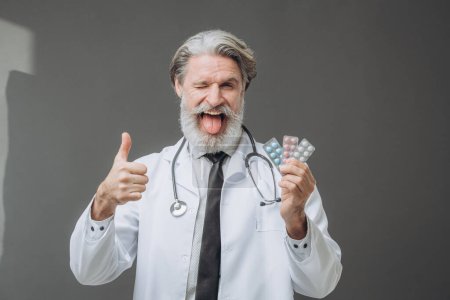 Photo for A man in a white medical coat is showing pills to the camera and holding a thumb up. - Royalty Free Image