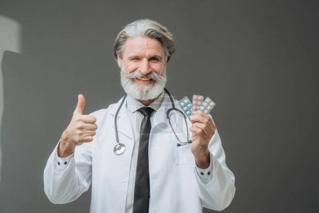 Photo for A man in a white medical coat is showing pills to the camera and holding a thumb up. - Royalty Free Image