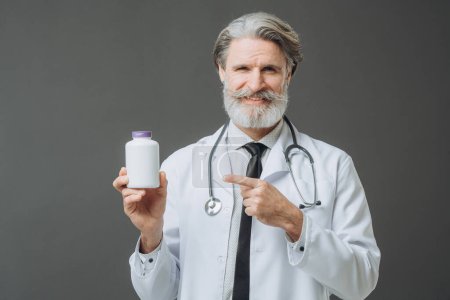 Photo for A gray-haired doctor in a white medical coat shows a bottle of pills to the camera. Medicine mockup. - Royalty Free Image