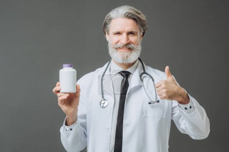 Photo for A man in a white medical coat is showing bootle of pills to the camera and holding a thumb up. - Royalty Free Image