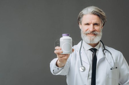 Photo for A gray-haired doctor in a white medical coat shows a bottle of pills to the camera. Medicine mockup. - Royalty Free Image