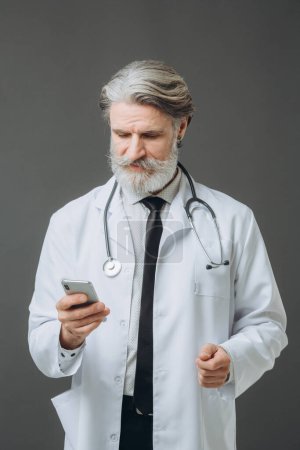 Photo for Portrait of a middle-aged male doctor with a phone in his hands on a gray background. - Royalty Free Image