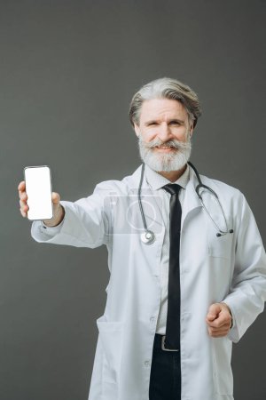 Téléchargez les photos : Un médecin aux cheveux gris en manteau médical blanc montre l'écran du téléphone à la caméra. Modèle de médicament. - en image libre de droit