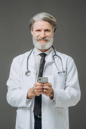 Photo for Portrait of a middle-aged male doctor with a phone in his hands on a gray background. - Royalty Free Image