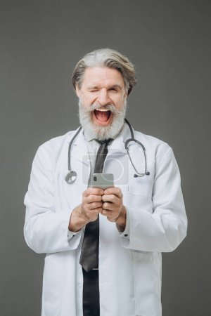 Photo for Emotional cheerful senior doctor with phone and hands on gray background. - Royalty Free Image