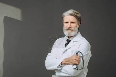 Photo for A senior doctor in a white coat with a phallendoscope in his hands poses on a gray background. The concept of healthcare and medicine. - Royalty Free Image