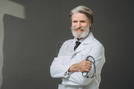 Photo for A senior doctor in a white coat with a phallendoscope in his hands poses on a gray background. The concept of healthcare and medicine. - Royalty Free Image