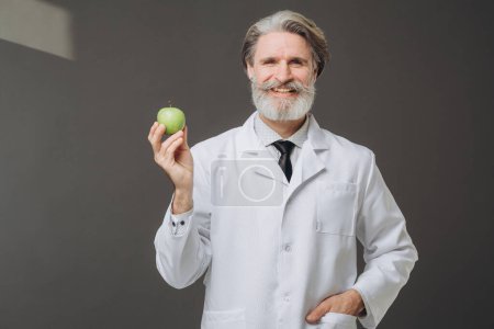 Schöner Oberarzt im weißen Mantel mit grünem Apfel in der Hand, der vor grauem Hintergrund posiert. Das Konzept der Gesundheitsfürsorge und Medizin.