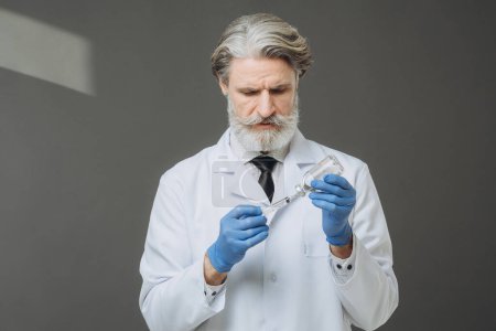 Photo for Gray-haired senior doctor in white coat holding syringe with injection, isolated on gray background. - Royalty Free Image