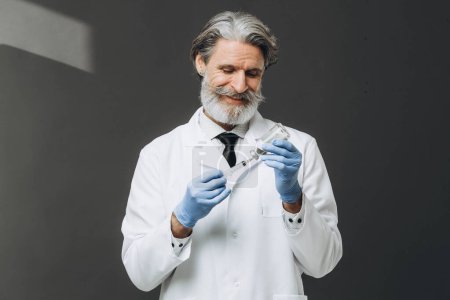 Médico senior de pelo gris de capa blanca que sostiene la jeringa con inyección, aislado sobre fondo gris.