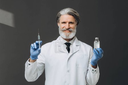 Photo for Gray-haired senior doctor in white coat holding syringe and bottle with injection, isolated on gray background. - Royalty Free Image