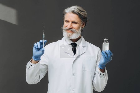 Médico senior de pelo gris de capa blanca que sostiene la jeringa y el frasco con inyección, aislado sobre fondo gris.