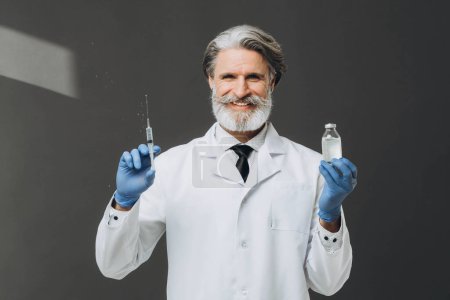 Photo for Gray-haired senior doctor in white coat holding syringe and bottle with injection, isolated on gray background. - Royalty Free Image