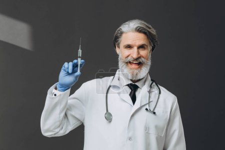 Photo for Portrait of a senior doctor in gloves and medical gown holding a syringe. isolated on gray background. - Royalty Free Image