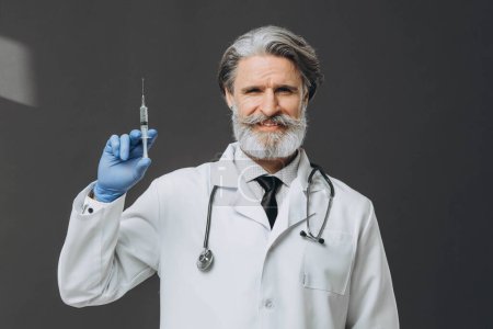 Photo for Portrait of a senior doctor in gloves and medical gown holding a syringe. isolated on gray background. - Royalty Free Image