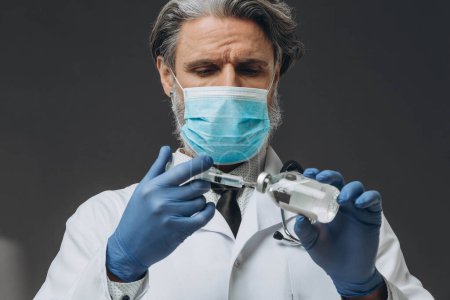 Photo for A senior doctor in a protective mask and gloves is preparing for an injection, drawing vaccine into a syringe. - Royalty Free Image