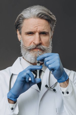 Foto de Retrato de un médico senior con guantes y bata médica sosteniendo una jeringa. aislado sobre fondo gris. - Imagen libre de derechos
