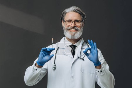 Photo for Senior doctor with grey hair holding syringe doing ''Ok'' sign with fingers, smiling friendly. - Royalty Free Image
