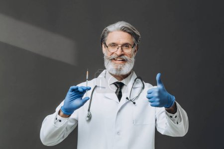 Photo for A senior doctor with gray hair is holding a syringe and giving a thumbs up, smiling warmly. - Royalty Free Image