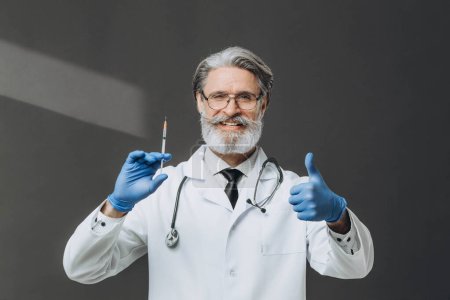 Photo for A senior doctor with gray hair is holding a syringe and giving a thumbs up, smiling warmly. - Royalty Free Image