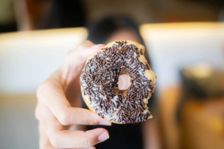 A person holding a bitten donut with chocolate sprinkles in front of the camera. The background is blurred.