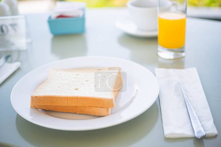 Two slices of white bread on a white plate, with a glass of orange juice and a cup in the background. The setting appears to be a breakfast table.