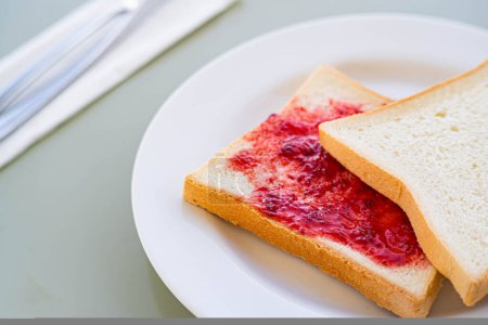 Close-up of two slices of white bread on a white plate, one with strawberry jam spread on it. A fork and knife are placed on a napkin in the background.