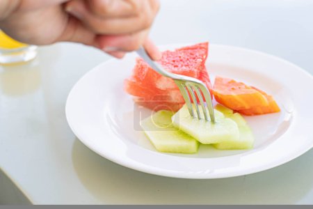 Téléchargez les photos : Une personne qui utilise une fourchette pour ramasser une tranche de melon dans une assiette blanche avec des tranches de fruits assorties, y compris la pastèque et la papaye. - en image libre de droit