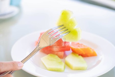 A close-up of a fork holding a piece of honeydew melon above a plate with slices of watermelon and honeydew melon.