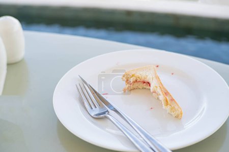 Ein teilweise verzehrtes Sandwich mit Marmelade auf einem weißen Teller, begleitet von Gabel und Messer, auf einem hellgrünen Tisch in der Nähe eines Pools.
