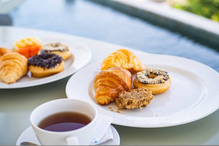A breakfast setting with a cup of tea, croissants, donuts, and a muffin on white plates near a pool.