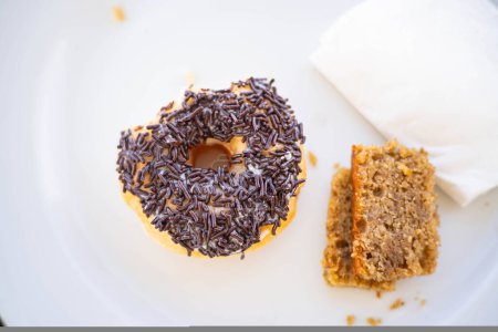 A donut with chocolate sprinkles and a piece of cake on a white plate, accompanied by a napkin.