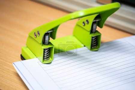 A green two-hole paper puncher being used on a stack of lined paper on a wooden desk.