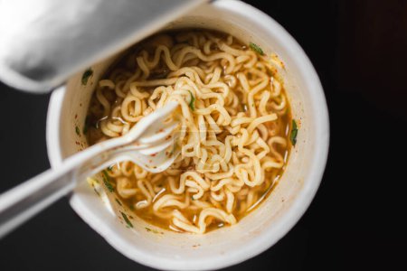 A cup of instant noodles with a plastic fork. The noodles are in a broth with pieces of meat and vegetables.