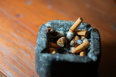 A close-up of a black ashtray filled with cigarette butts on a wooden table.