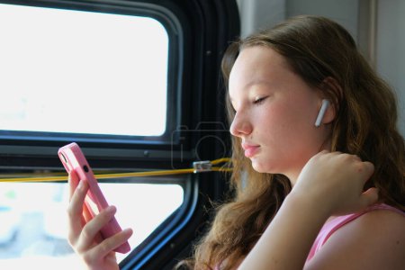 The girl puts on headphones, listens to audio messages and music, listens to songs and podcasts on an online application. Language learning on public transport. Keeping a teenager quiet in a public