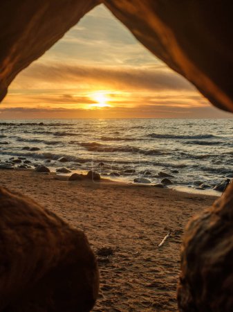Peering out from the depths of a cave at Veczemju Klintis Beach, the sunset casts its radiant glow over the horizon, painting the sky in hues of gold and orange.