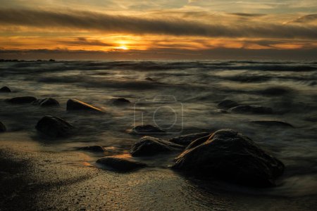 Téléchargez les photos : Dans l'étreinte tranquille de Veczemju Klintis, le soleil couchant peint une scène envoûtante sur la plage rocheuse, capturée dans une beauté éthérée grâce à une longue exposition - en image libre de droit