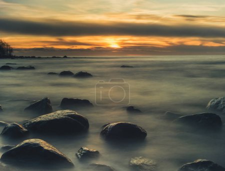 As daylight fades, Veczemju Klintis beach transforms into a realm of timeless beauty, where the rhythmic waves and golden hues of sunset intertwine in a captivating long exposure shot