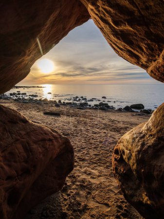 Peering out from the depths of Veczemju Klintis Cave, the silhouette of twilight unveils a mesmerizing vista as the sun bids farewell, casting its radiant glow upon the rocky beach