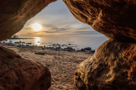 Eingebettet in die schützenden Arme der Höhle Veczemju Klintis erleben wir den faszinierenden Tanz der Farben des Sonnenuntergangs, der den felsigen Strand mit Wärme und Staunen bemalt.