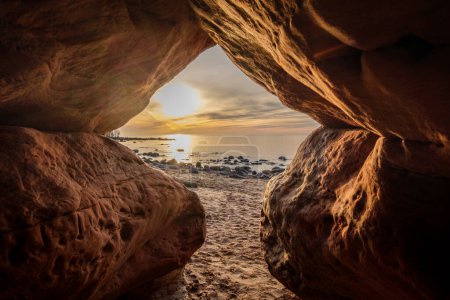 Within the depths of Veczemju Klintis Cave, twilight descends like a gentle melody, enveloping the rocky beach in a surreal tapestry of sunset magic.