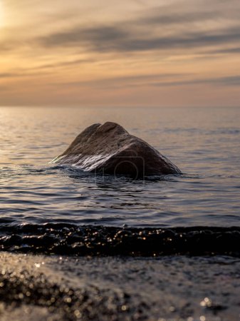 As the sun sets on Veczemju Klintis, a lone stone stands silently amidst the gentle waves, reflecting the colors of twilight in tranquil serenity.