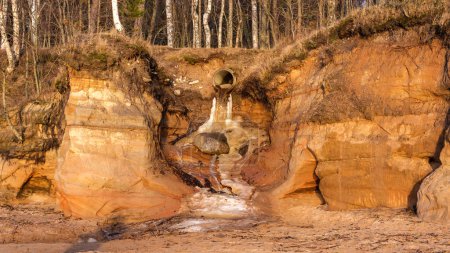 In the golden glow of sunset, water pours gracefully from a weathered pipe onto the washed coastline of Veczemju Klintis, creating a serene and picturesque coastal waterfall scene