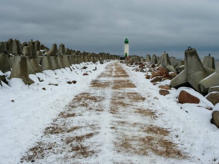 Spuren markieren den verschneiten Weg zum Südpier von Ventspils, wo das Licht des Leuchtturms durch die kalte Winterluft dringt.