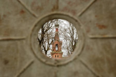 Téléchargez les photos : Par l'ouverture rustique d'une clôture métallique, le clocher de l'église se dresse haut, symbole de la foi et du patrimoine à Kuldiga, Latvija - en image libre de droit